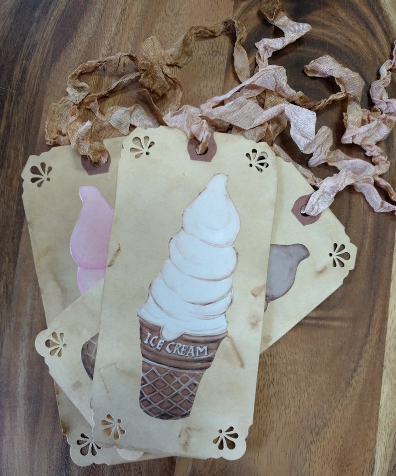 May include: Four brown paper gift tags with scalloped edges. Each tag has a different image of a dessert: a pink ice cream cone, a white ice cream cone with the text "ICE CREAM", a brown ice cream cone, and a brown bird. The tags are tied together with brown and pink ribbon.