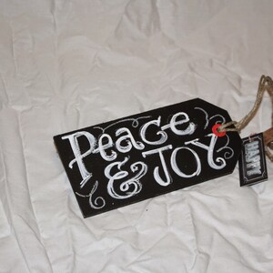 May include: Black chalkboard tag with white lettering that reads "Peace & Joy". The tag is tied with twine and has a small brown tag that reads "Happy".