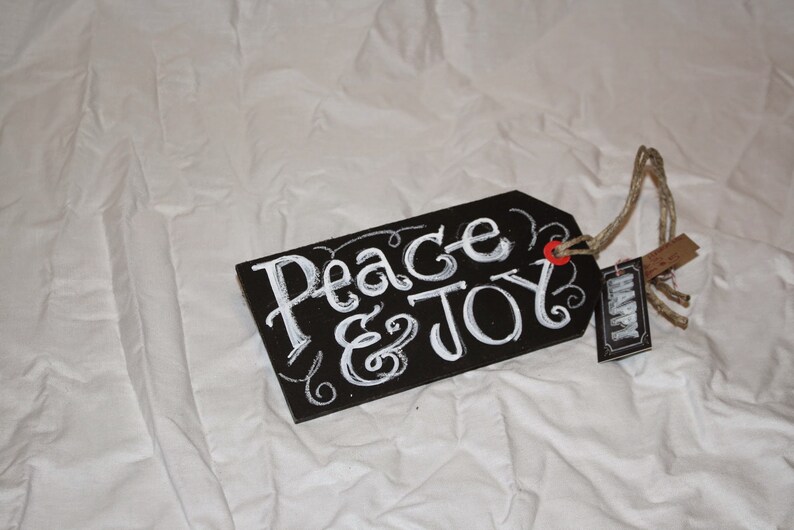 May include: Black chalkboard tag with white lettering that reads "Peace & Joy". The tag is tied with twine and has a small brown tag that reads "Happy".