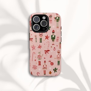 May include: A pink phone case featuring a repeating pattern of Christmas-themed illustrations. The design includes nutcrackers, candy canes, stars, gift boxes, and snowflakes in red, green, and white. The case is designed to fit a smartphone.