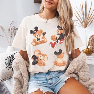 May include: Cream-colored t-shirt featuring four cartoon teddy bears with black Mickey Mouse ears. One bear holds a balloon, another has the letter "M" on its chest. The bears are in various poses, adding a playful touch to the shirt.