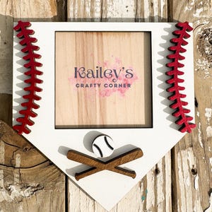 May include: White wooden baseball home plate picture frame with red stitching and a baseball and crossed bats. The frame has a wood background with the text "Kailey's Crafty Corner".