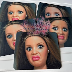 May include: A set of five square coasters featuring a close-up of a doll's face with exaggerated expressions. The doll has brown hair, blue eyes, and pink lipstick. The coasters are arranged on a white surface.