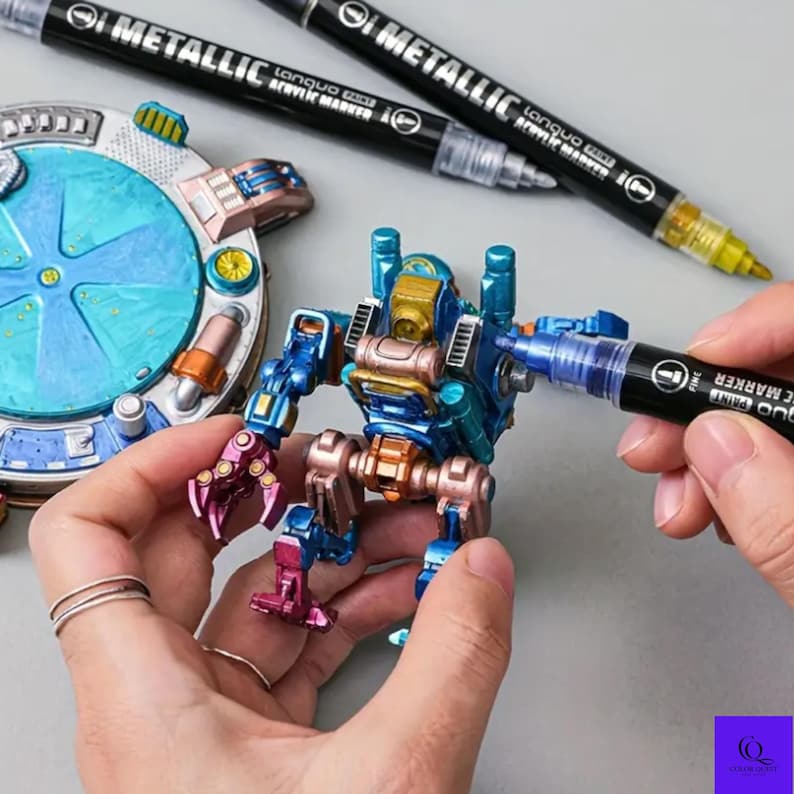 May include: A person using a metallic acrylic marker to paint a small, colorful robot figurine. The robot is blue, gold, and pink. Two additional markers are in the background. The markers have the text "METALLIC" and "lanquo ACRYLIC MARKER".