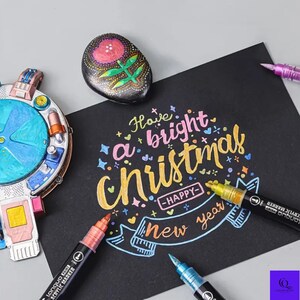May include: A black card with the words "Have a bright Christmas - Happy New Year" in colorful lettering. Several acrylic markers in various colors are arranged around the card. A painted rock with a floral design is also present.