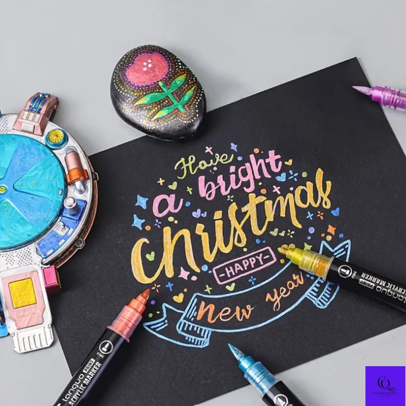 May include: A black card with the words "Have a bright Christmas - Happy New Year" in colorful lettering. Several acrylic markers in various colors are arranged around the card. A painted rock with a floral design is also present.