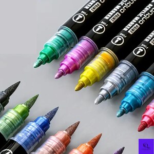 May include: A collection of metallic acrylic paint markers in various colors, including green, pink, yellow, silver, and blue. The markers have black barrels with white text and fine tips, ideal for art projects and crafts.