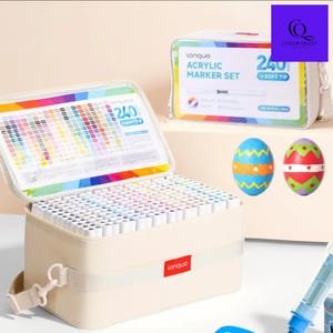 May include: A beige acrylic marker set with 240 soft tip markers, displayed in an open case. The case has a rainbow-colored lid and a color chart. A second case is visible, along with two decorated Easter eggs.
