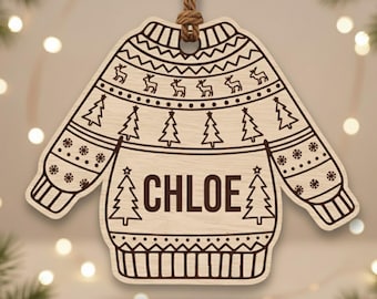 Könnte beinhalten: Weihnachtsornament aus Holz in Form eines Pullovers mit dem Namen "CHLOE" in der Mitte. Das Pullover-Design umfasst Bäume, Rentiere und Schneeflockenmuster. Ein brauner Faden ist zum Aufhängen befestigt.