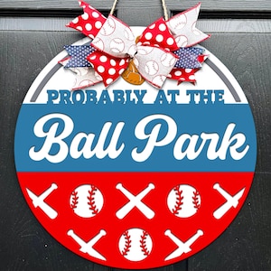 May include: A circular wooden sign with a baseball theme. The sign has the text "Probably at the Ball Park" in white. It's decorated with baseballs, bats, and a red, white, and blue bow, perfect for a door.