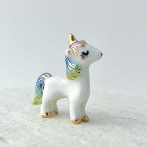May include: A small, white ceramic unicorn figurine with a gold horn, hooves, and accents. The mane and tail feature pastel colors including blue, green, and pink. The unicorn has a delicate, hand-painted appearance.