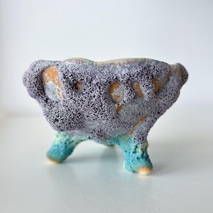 May include: A small, three-legged ceramic bowl featuring a highly textured, porous surface. The upper portion is finished in a mottled lavender and grey glaze, while the three short legs transition into a vibrant, bright turquoise blue hue.