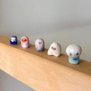 May include: A collection of five small, ceramic figurines with rounded shapes and painted faces. The figurines are in shades of blue, pink, and white, with delicate details. They are arranged on a wooden surface, creating a whimsical display.