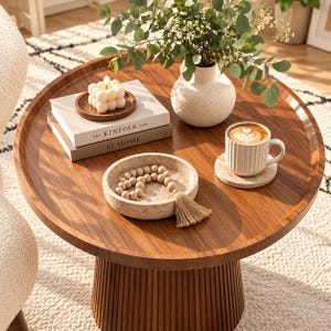 May include: A round wooden coffee table with a fluted pedestal base. On the surface are two stacked books titled The Kinfolk Home and At Home, a white bubble candle, a speckled vase with greenery, a stone bowl with wooden beads, and a coffee mug.