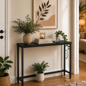 May include: A rectangular black metal console table with four thin legs and a horizontal support bar. The table surface holds a ribbed beige vase with greenery, a glass candle holder, and a stack of three books titled Kinfolk, This Is Home, and Live Beautifully.