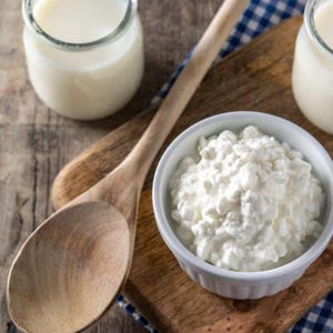 Puede incluir: Una cuchara de madera descansa junto a un pequeño cuenco blanco lleno de requesón. También hay dos tarros de cristal llenos de un líquido blanco. Los artículos están colocados sobre una tabla y una mesa de madera.