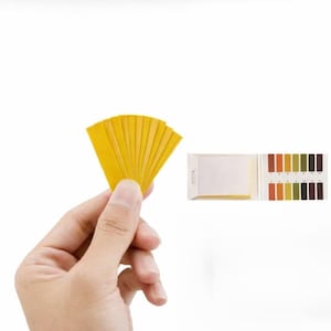 Puede incluir: Un conjunto de tiras reactivas de pH amarillas dispuestas en abanico, acompañadas de un pequeño folleto con una tabla de referencia codificada por colores con valores numerados del 1 al 14 para medir la acidez o alcalinidad de un líquido.
