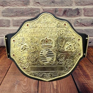 May include: A gold-colored wrestling championship belt with the words "WORLD HEAVYWEIGHT WRESTLING CHAMPION" engraved on it. The belt has a black leather strap and is decorated with red jewels and ornate designs.