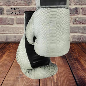 Pode incluir: Um par de luvas de boxe com um padrão texturizado semelhante a pele de cobra em tons de branco e cinza. As luvas têm detalhes em preto e uma alça de pulso. O fundo apresenta uma parede de tijolos e tábuas de madeira.