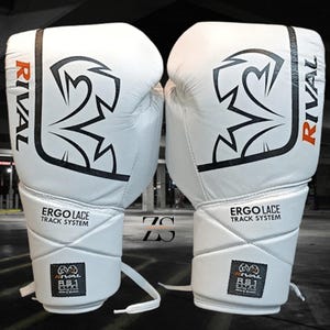 Puede incluir: Un par de guantes de boxeo blancos con detalles en negro y naranja. Los guantes presentan el logotipo "RIVAL" y el texto "ERGO LACE TRACK SYSTEM". Estos guantes están diseñados para boxeo y deportes de combate.
