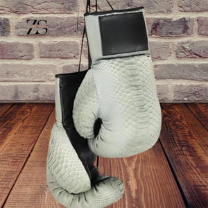 Puede incluir: Un par de guantes de boxeo en un patrón de piel de serpiente gris claro con detalles en negro. Los guantes cuelgan contra una pared de ladrillo y una superficie de madera. Los guantes tienen una apariencia texturizada y están diseñados para boxeo o entrenamiento.