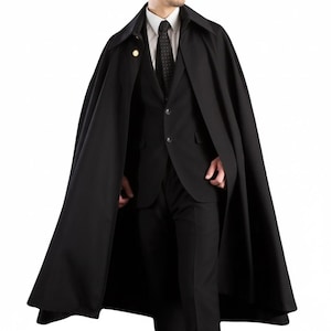 May include: A black cape with a wide collar and a gold button detail. The cape is worn over a black suit, white shirt, and black tie. The cape is a classic style, suitable for formal occasions.