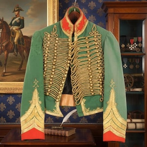 Può includere: Una giacca militare verde con finiture e bottoni dorati, colletto e polsini rossi e dettagli decorativi in corda dorata. La giacca è appesa a un gancio. Un dipinto di un soldato a cavallo è sullo sfondo.