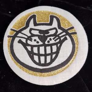 May include: A round, white patch featuring a cartoon cat face with a wide grin and large teeth, outlined in black. The cat is surrounded by a gold glitter border. The patch is likely made of fabric.