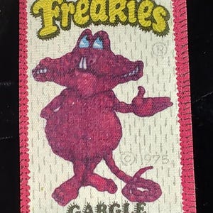 May include: A vintage Freakees sticker featuring a purple cartoon alligator named Gargle. The alligator has blue eyes and is standing on two legs with one arm raised. The sticker has a white background with a pink border and the text "Freakies" in yellow.