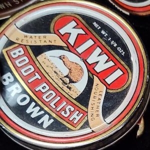 May include: A close-up of a can of Kiwi Boot Polish in brown. The label features the word "KIWI" in white on a red background, a kiwi bird illustration, and the text "WATER RESISTANT" and "LEATHER NOURISHING".