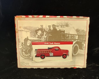 May include: A red 1948 Task Master fire truck model displayed in a box with a vintage photograph of firefighters. The model truck has a ladder and silver accents. The box has red and white striped edges.