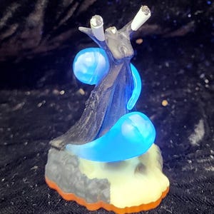 May include: A fantasy figurine featuring a dark, tree-like figure with glowing blue orbs and accents. The base is a mix of gray, white, and orange. The figure has white tips and a dark, textured appearance.