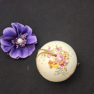 May include: A cream-colored, spherical decorative object with floral designs in pink, yellow, and blue, and small perforations. A purple flower with a pearl center is next to it. A gold string is attached to the sphere.