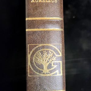 May include: A brown hardcover book with gold lettering. The spine features the names "PLATO", "EPICTETUS", "MARCUS AURELIUS", and a gold emblem of a tree within a "G". The book is part of "THE HARVARD" series.