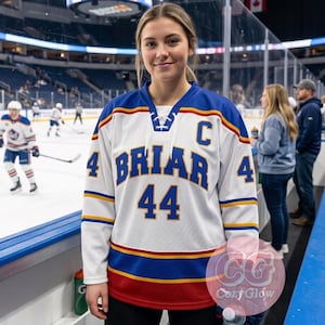 Op de afbeelding: Een wit hockey-shirt met blauwe en gouden accenten. Het shirt toont de naam "BRIAR" en het nummer "44" in blauw. Een persoon draagt het shirt in een ijshockeyarena.