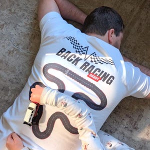 Puede incluir: Una camiseta blanca con un diseño de circuito de carreras y las palabras "BACK RACING DADDY" impresas en la espalda. Un niño sostiene un coche de juguete en la camiseta. La camiseta tiene un gráfico de bandera a cuadros.