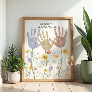 Könnte beinhalten: Ein gerahmter Druck mit einem hellen Holzrahmen. Das Kunstwerk zeigt drei Handabdrücke in Lila, Gelb und Braun mit den Worten "WE LOVE YOU grandma" darüber. Darunter befinden sich Aquarellblumen in verschiedenen Farben.