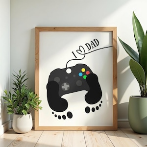 May include: Framed print featuring a black video game controller design with colorful buttons and a heart-shaped "I ♡ DAD" text. The controller is integrated with black footprint shapes. The artwork is set in a light brown wooden frame.