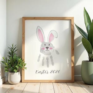 May include: A framed print featuring a handprint bunny design with pink inner ears and whiskers. The bunny's body is formed by a handprint. The text "Easter 2026" is written in a script font below the bunny. The frame is made of light-colored wood.