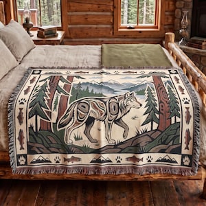 Peut inclure: Un plaid tissé présentant une illustration centrale de loup dans un décor forestier avec des montagnes en arrière-plan. La bordure affiche un motif répétitif de poissons, d'empreintes de pattes et de silhouettes de montagnes en tons brun, vert et crème.