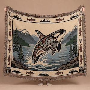 May include: A woven throw blanket featuring a central graphic of an orca breaching water, surrounded by a landscape of mountains and pine trees. The border displays a repeating pattern of fish and mountain peaks, finished with a fringed edge.