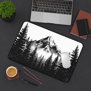 Puede incluir: Alfombrilla de ratón en blanco y negro con un diseño de montaña y bosque. El diseño muestra una ilustración detallada de una cordillera con árboles en primer plano. Un ratón de ordenador blanco está en el lado derecho.