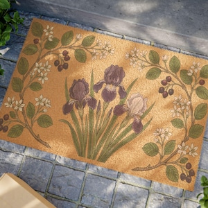 Può includere: Un zerbino rettangolare con un design floreale. Il tappeto presenta tre iris viola al centro, circondate da un bordo di foglie verdi, fiori bianchi e bacche scure su uno sfondo beige. Il tappeto è realizzato in un materiale di fibra naturale.