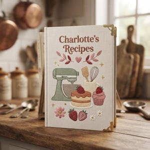 May include: A cream-colored fabric-covered recipe book titled Charlotte's Recipes with gold-tone metal corner protectors. The cover features embroidered illustrations of a stand mixer, a whisk, a spatula, a cake, a cupcake, and strawberries.