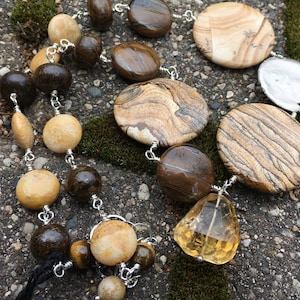 Pode incluir: Um colar longo feito com pedras marrons, bege e brancas. O colar tem uma variedade de formas e tamanhos de pedras, incluindo redondas, ovais e retangulares. As pedras são enfiadas em uma corrente de prata.