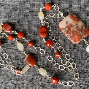 Op de afbeelding: Een zilveren ketting met een grote hanger van rode en witte jaspis. De ketting heeft ook kleinere rode jaspis kralen en witte kralen.