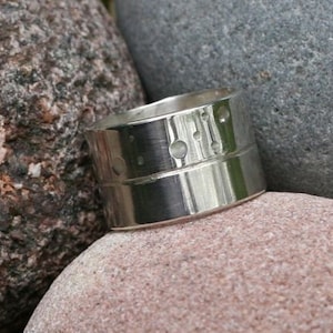 Può includere: Un anello d'argento con una superficie testurizzata e una fascia di piccoli cerchi. L'anello è appoggiato su una roccia.