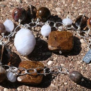 Könnte beinhalten: Ein silbernes Kettenarmband mit braunen und weißen Achatperlen. Das Armband hat einen silbernen Verschluss mit einem kleinen Anhänger, auf dem "S&J" steht.