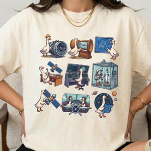 May include: Ivory-colored t-shirt featuring a series of cartoon geese engaged in various activities related to space and aviation. The illustrations include geese operating machinery, using computers, and exploring space. The shirt has a crew neck.