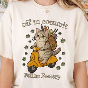 May include: Cream-colored t-shirt with a graphic of a gray tabby cat riding a yellow scooter, carrying a backpack. The text "off to commit Feline Foolery" is above and below the cat. Green paw prints surround the cat.